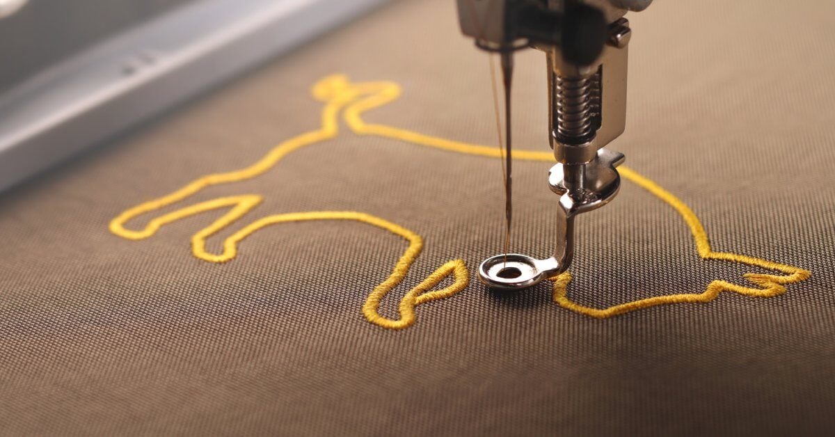 A close-up shot of a machine embroidery needle quickly stitching a golden bull pattern into brown, stabilized fabric.